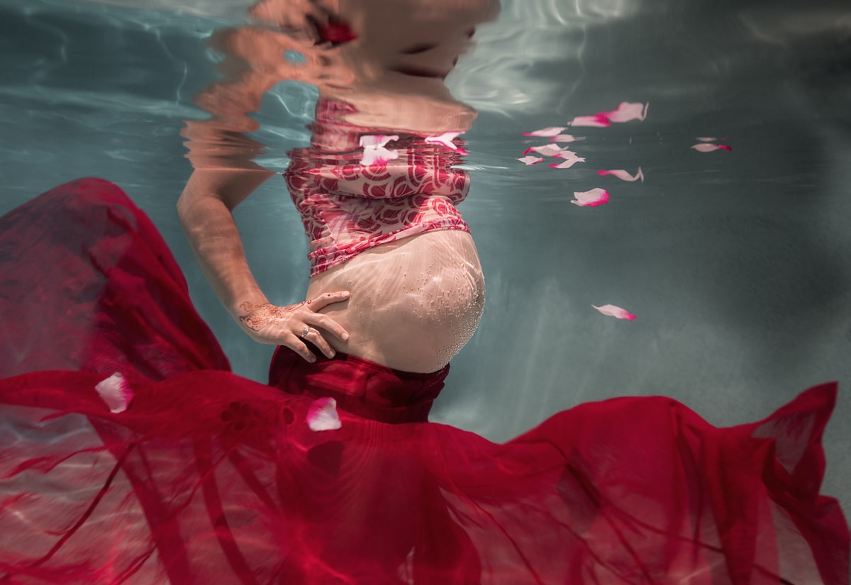 Pregnant belly underwater with bubbles on skin and floating flower petals and red fabric and henna tattoo on hand